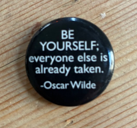 A black button pin displaying the quote: 'BE YOURSELF; everyone else is already taken.' by Oscar Wilde, resting on a wooden surface.