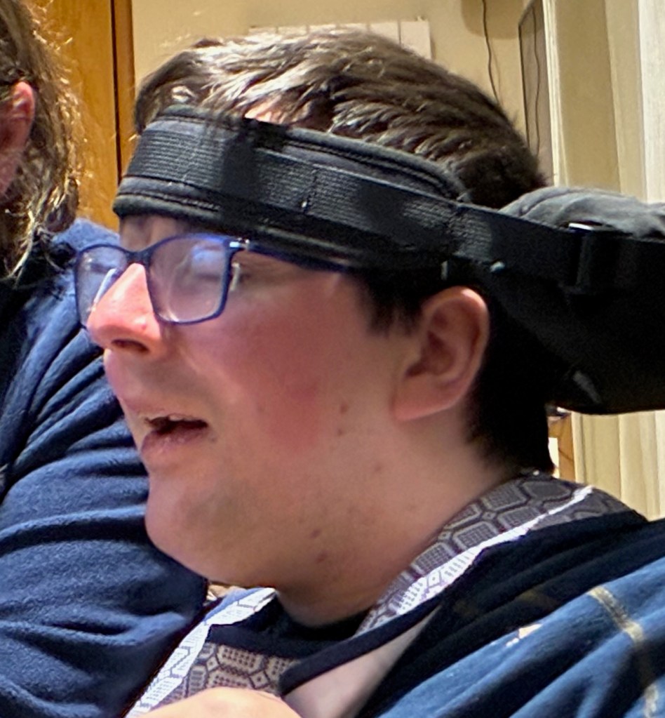 A close-up of a young man with glasses and a headband, looking to the side with an expression of concentration. He is wearing a dark-colored shirt and there are two people visible behind him.