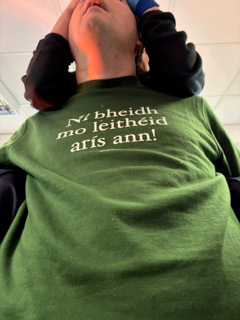 A person wearing a green shirt with the text 'Ná beidh mo leithéid arís ann!' displayed prominently on the front. The individual is tilting their head back, while another person is partially visible in the background.