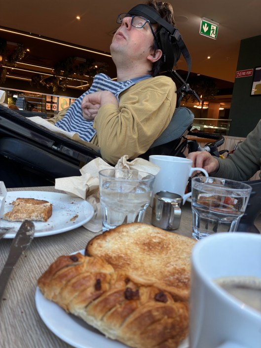 A breakfast scene featuring a person in a wheelchair looking up, partially out of focus. In the foreground, there are plates of toast and a croissant, along with cups of coffee and glasses of water on a wooden table.