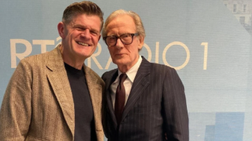 Two men posing together for a photograph, smiling at the camera. The man on the left is wearing a casual outfit with a light jacket, while the man on the right is dressed in a formal suit and glasses. The background features the words 'RTÉ RADIO 1' on a light blue backdrop.