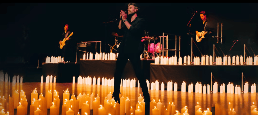 A singer performing on stage surrounded by lit candles, with a band visible in the background.